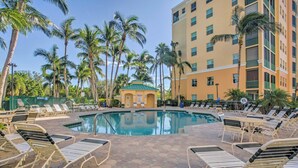 A pool area with lounge chairs and a small building, surrounded by palm trees and a multi-story building. - Waterfront Punta Gorda Condo w/ Pool Access! (Punta Gorda)