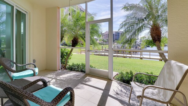 A balcony with wicker chairs, a ceiling fan, and a view of a grassy area with palm trees and buildings. - Waterfront Punta Gorda Condo w/ Pool Access! (Punta Gorda)