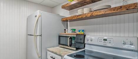 Une cuisine compacte avec un réfrigérateur blanc, un four à micro-ondes et une cuisinière. Des étagères en bois, situées au-dessus de la cuisinière, servent à ranger la vaisselle et une bouteille.