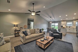 A spacious living room with a sofa, coffee table, and a ceiling fan. A kitchen with white cabinets and a dining area with a table and chairs are visible in the background.