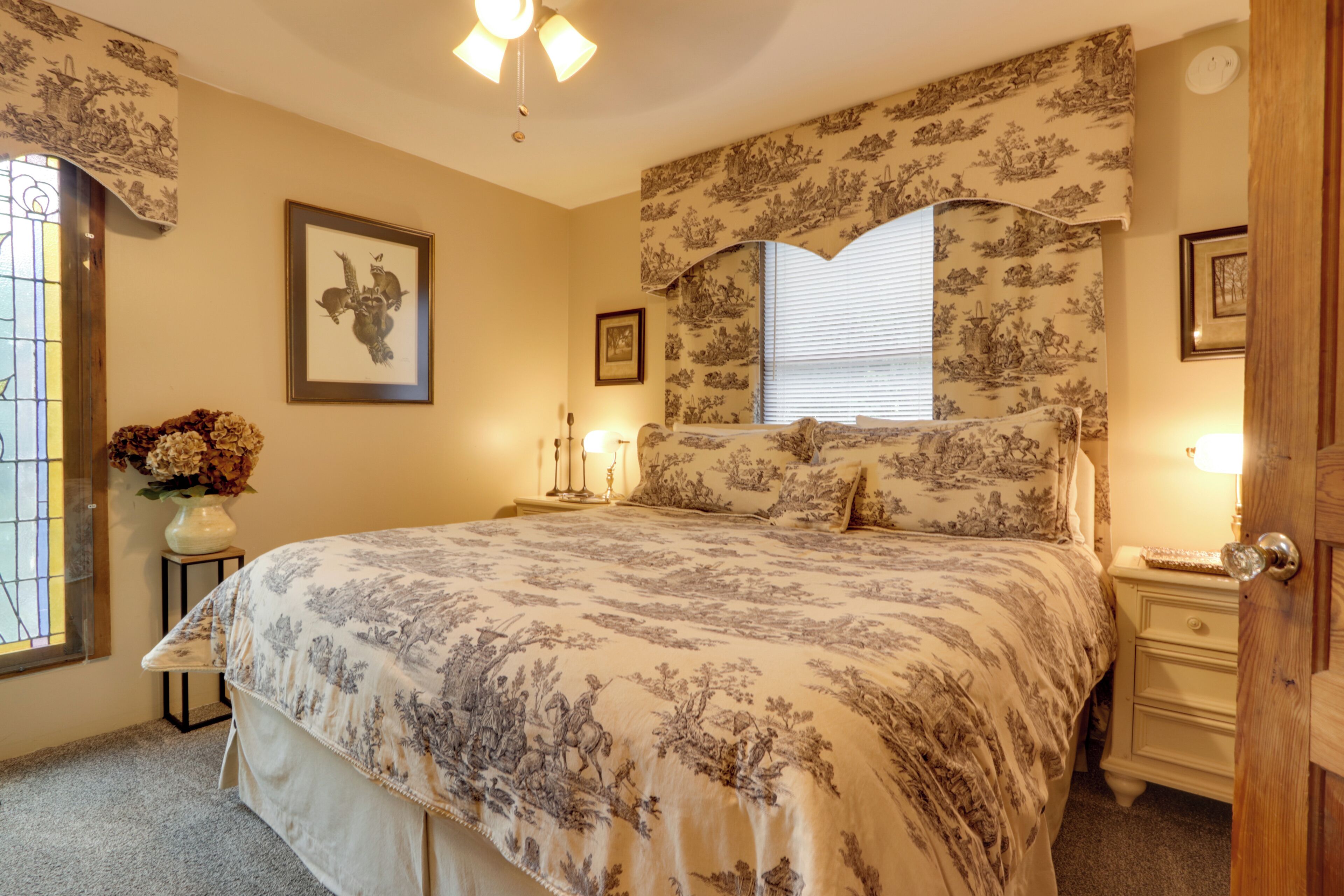 A bedroom with a large bed, floral wallpaper, a framed picture on the wall, a vase with flowers, and a window with blinds.