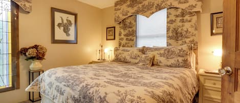 A bedroom with a large bed, floral wallpaper, a framed picture on the wall, a vase with flowers, and a window with blinds.