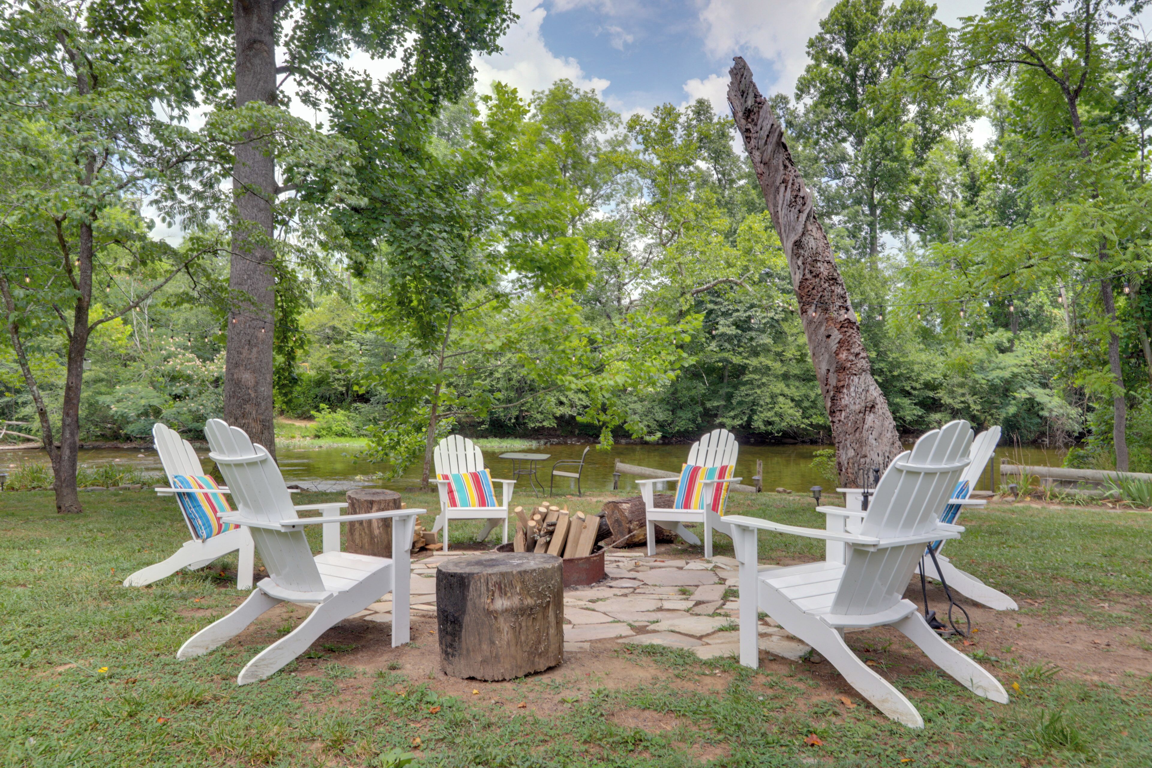 A serene outdoor seating area with Adirondack chairs and a fire pit surrounded by lush greenery and a pond.
