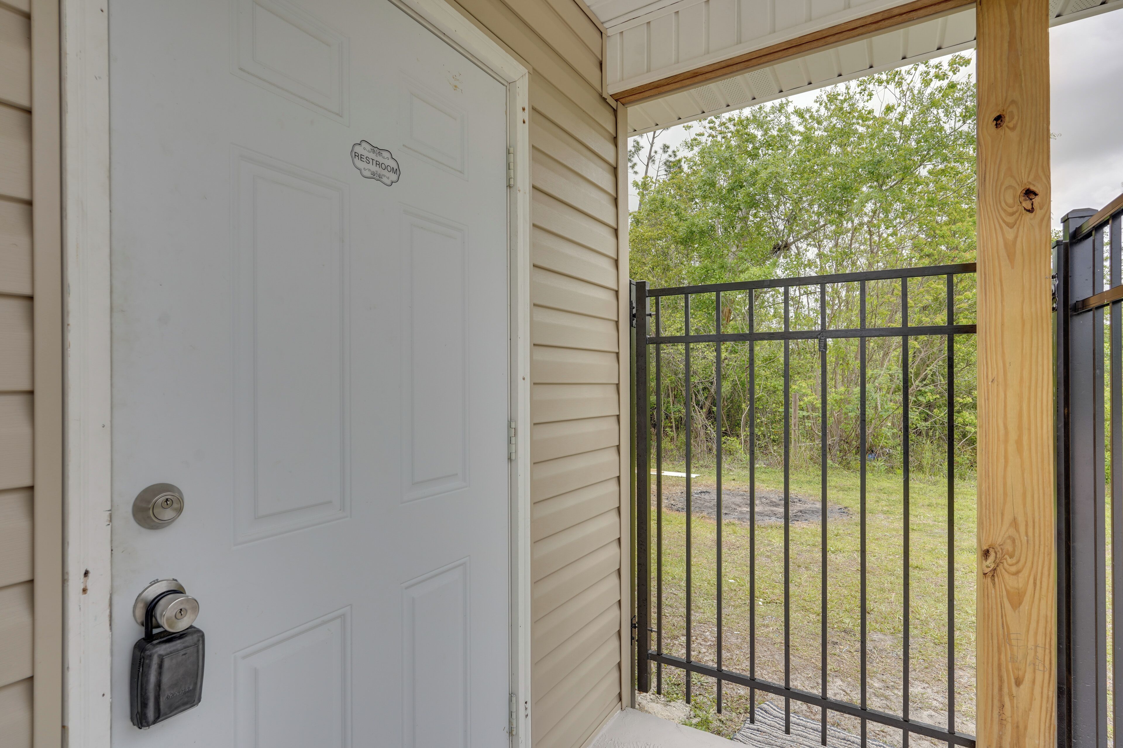 A white door with a keyhole and a lock, a small sign above it, and a black metal gate leading to a grassy area with trees.