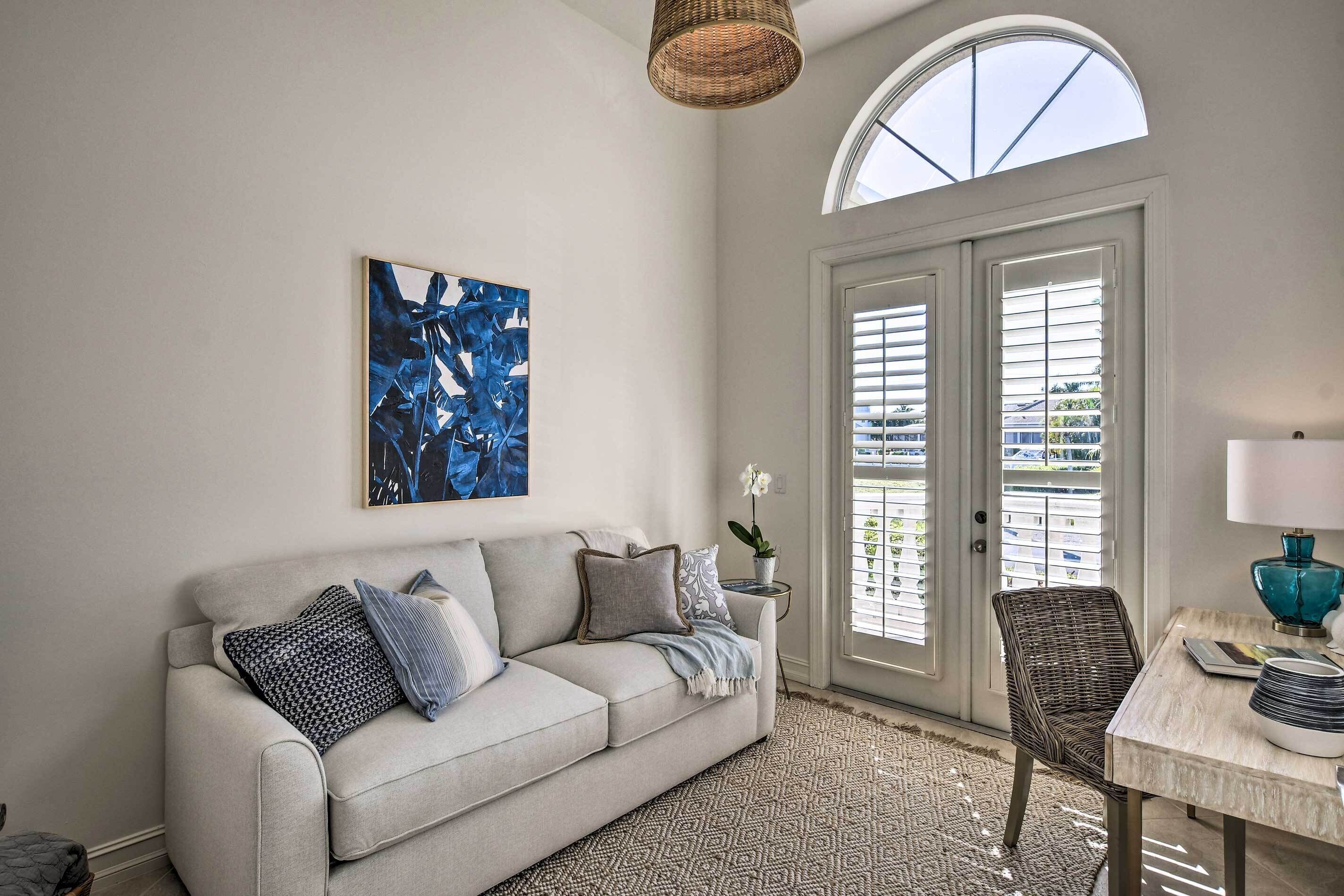 A living room with a sofa, a table with a lamp, a wicker chair, and a large window with shutters.