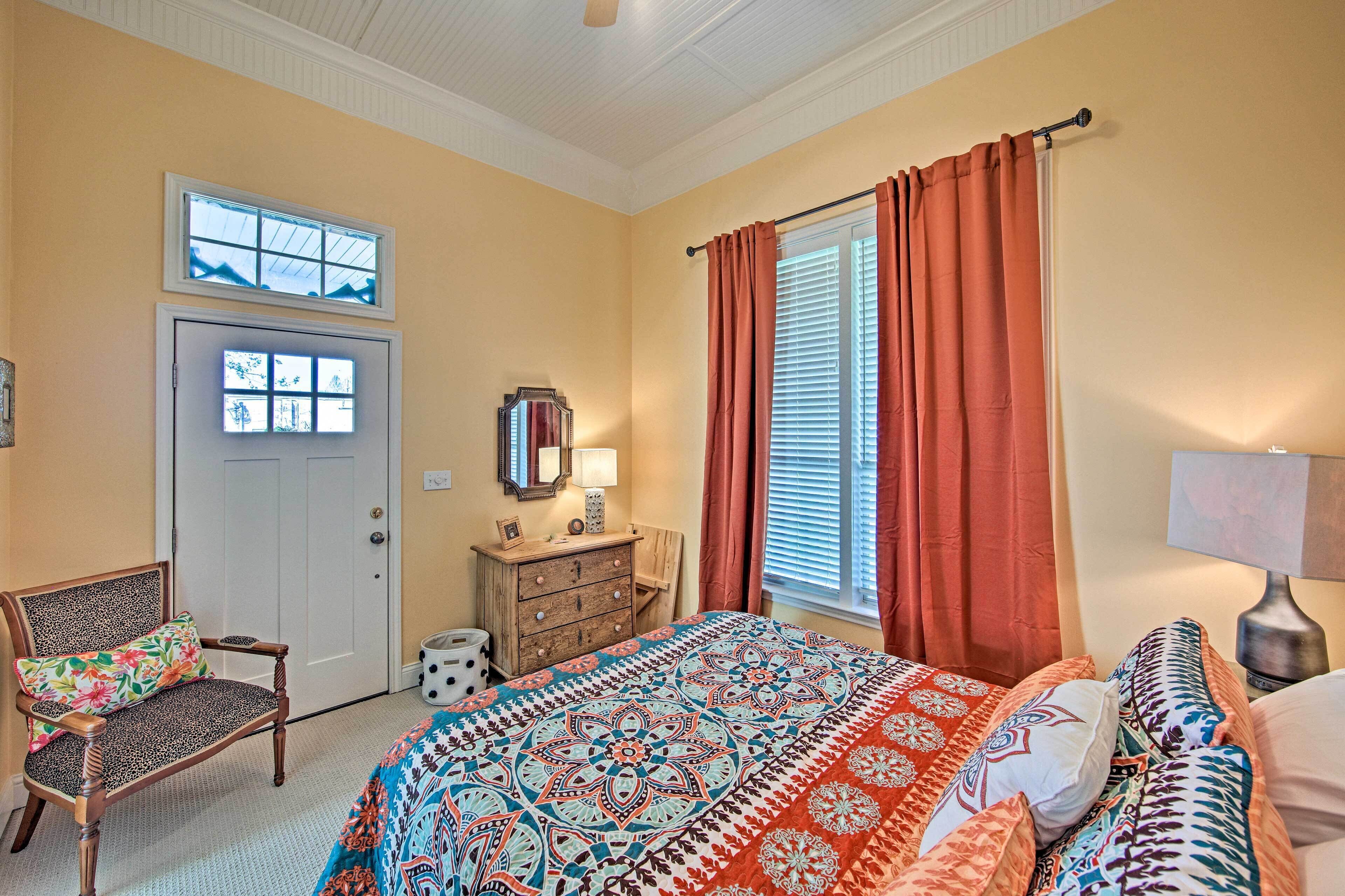 A bedroom with a bed, a chair, a dresser, a window with curtains, and a door with a window above it.