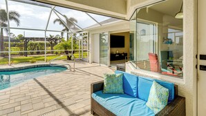 Área da piscina com um alpendre envidraçado, um sofá de vime com almofadas e vista para a piscina e vegetação.