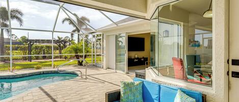A poolside area with a glass-enclosed patio, a wicker sofa with cushions, and a view of the pool and greenery.