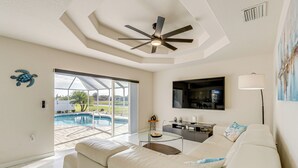 Uma sala de estar moderna com um sofá branco, uma mesa de centro de vidro, um ventilador de teto, uma TV e vista para uma piscina através de portas de vidro.