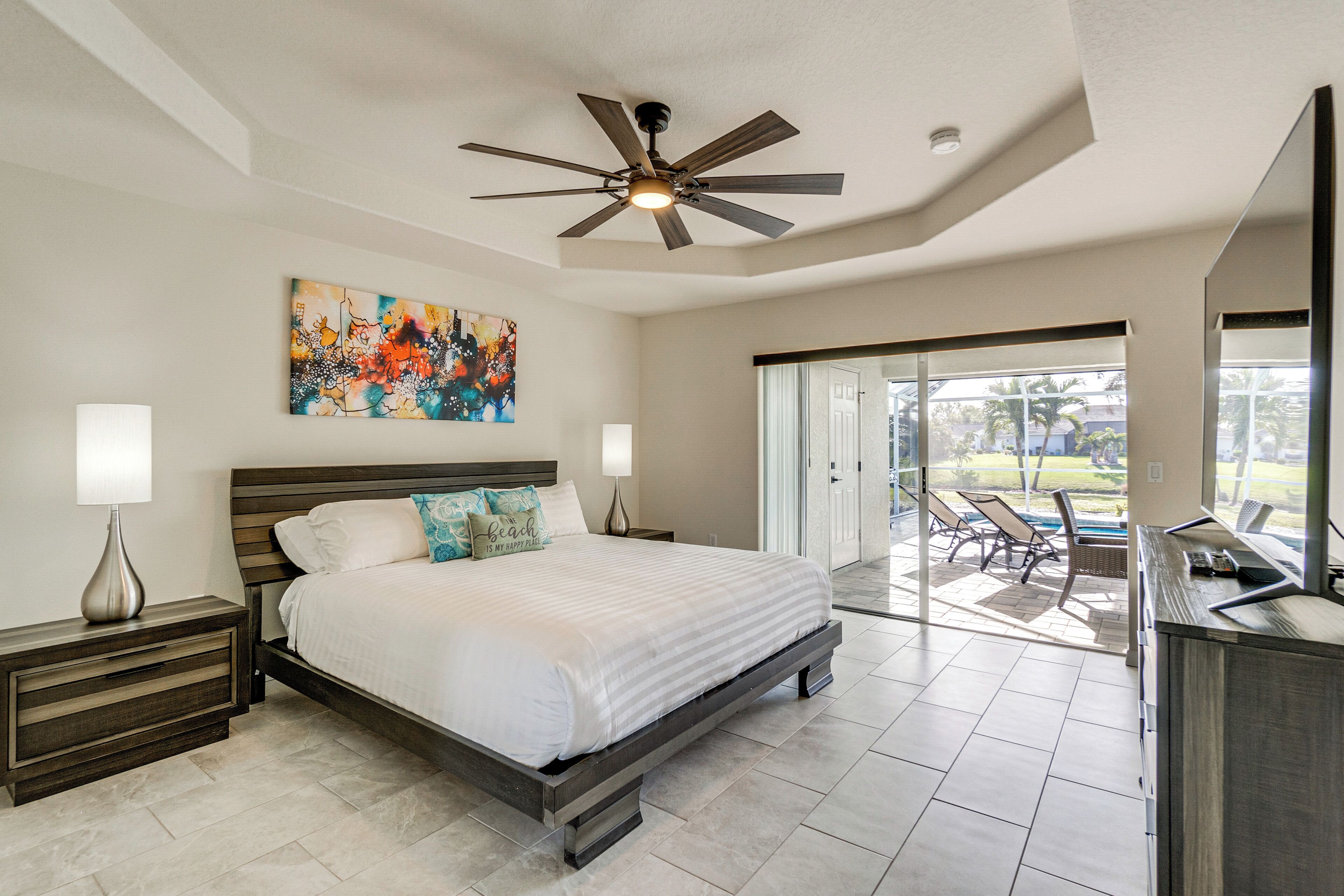 A bedroom with a large bed, a ceiling fan, a painting, and a view of the outside.