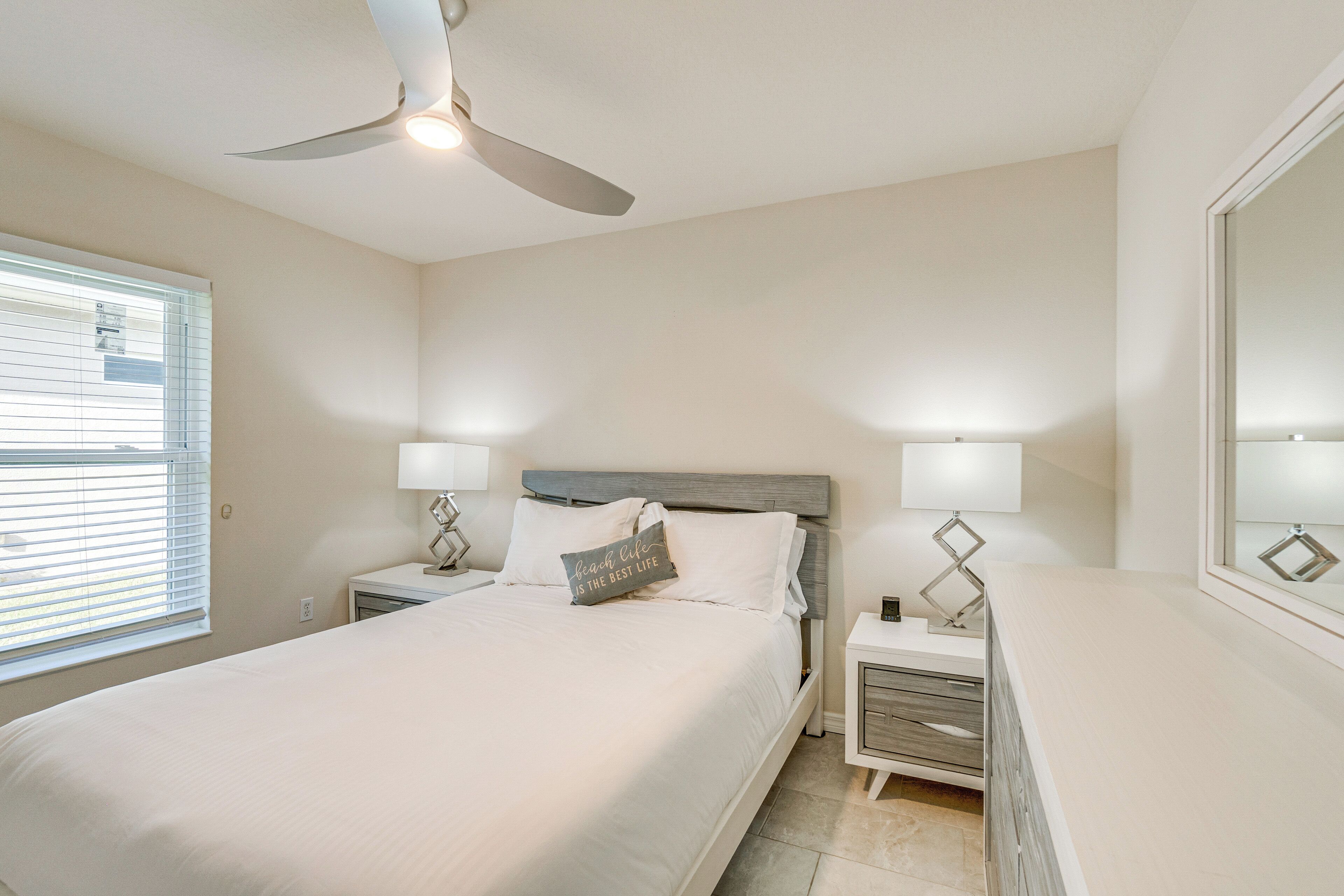 A bedroom with a bed, two bedside tables with lamps, a ceiling fan, a window with blinds, and a mirror on the wall.