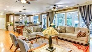Ein großzügiges Wohnzimmer mit einer Couch, Sesseln, einem Couchtisch und einer Lampe. Es gibt Deckenventilatoren, eine Küche im Hintergrund und große Fenster mit Vorhängen.