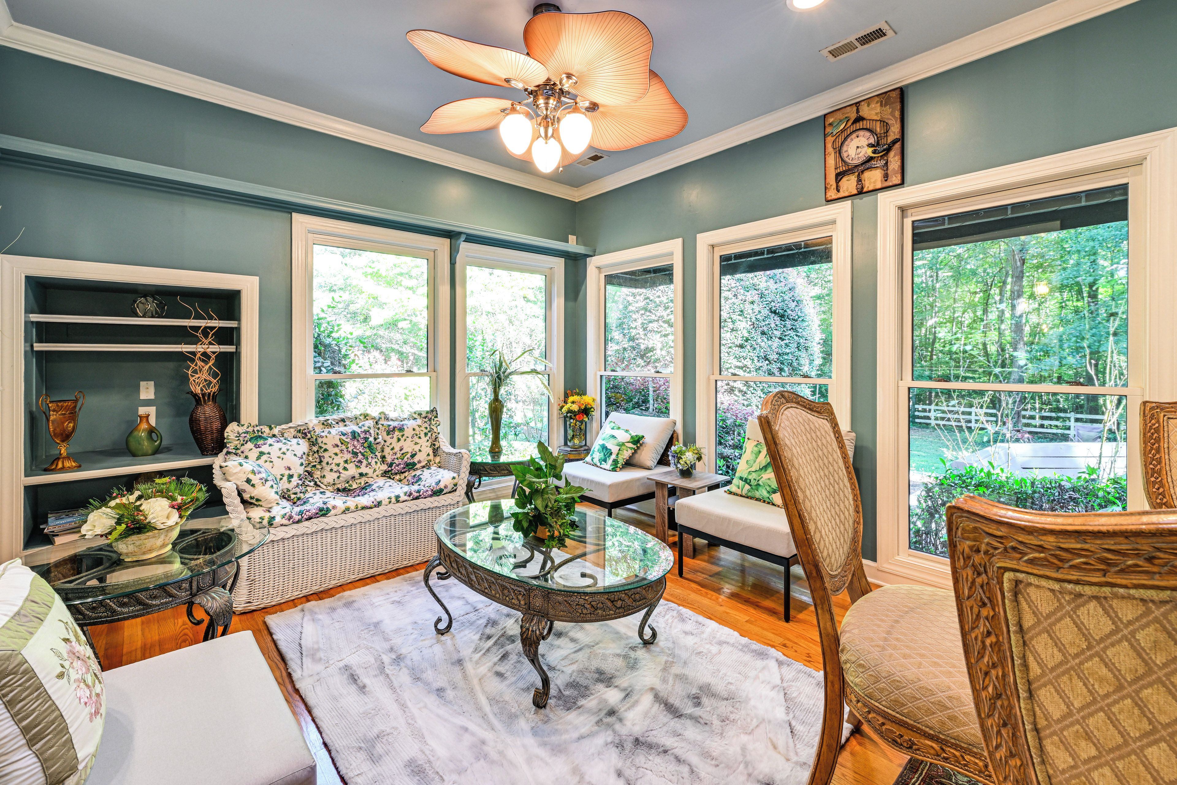 A spacious room with a ceiling fan, large windows, a sofa, a chair, and a table with a vase.