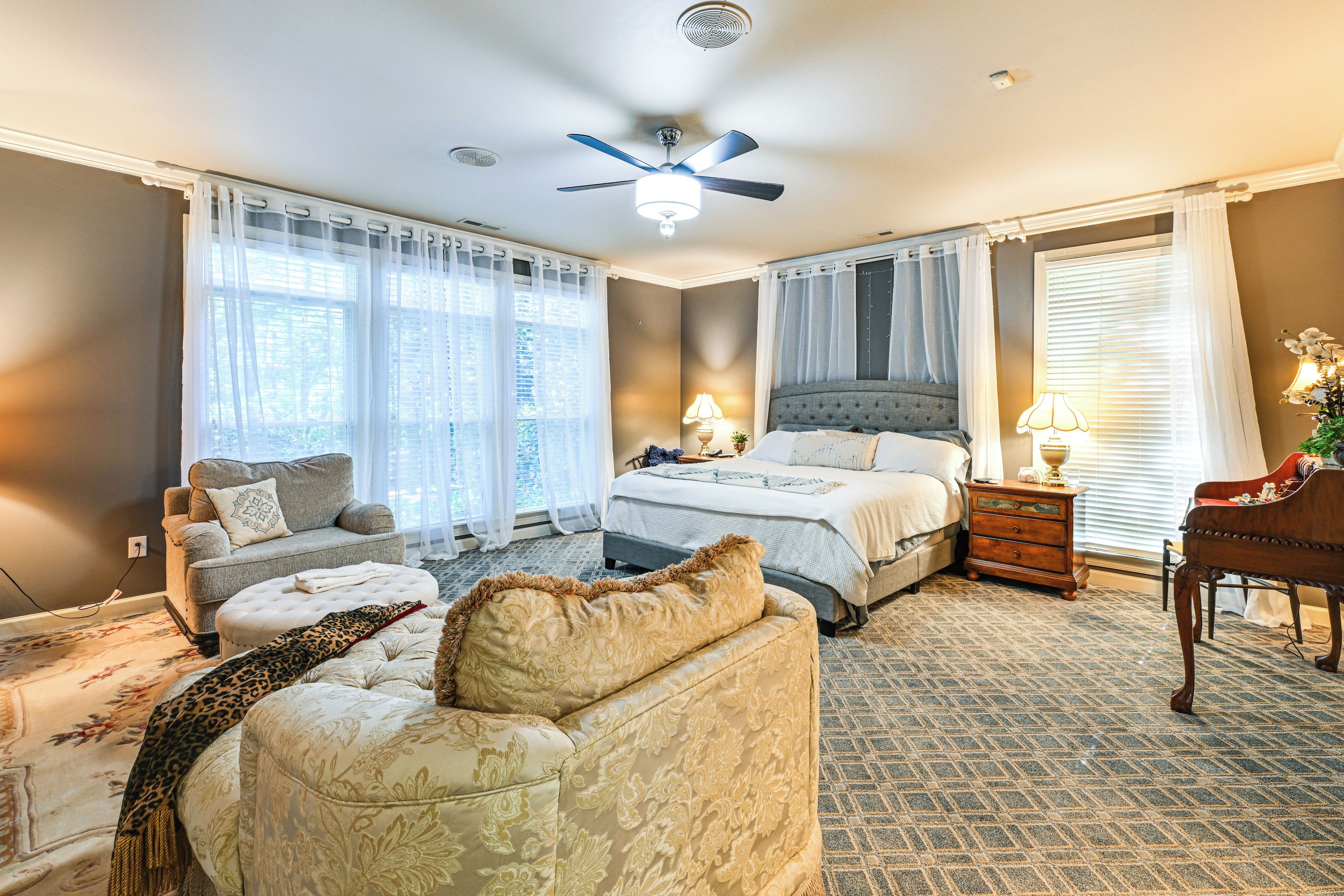 A bedroom with a bed, two bedside tables, a nightstand, a chair, a sofa, a ceiling fan, and a window with curtains.