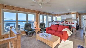 A spacious living room with a red sofa, black leather chairs, and a wooden coffee table. A ceiling fan is visible, and large windows offer a view of the ocean. - Exquisite Oceanside House w/ Pacific Views & Deck! (Tillamook)