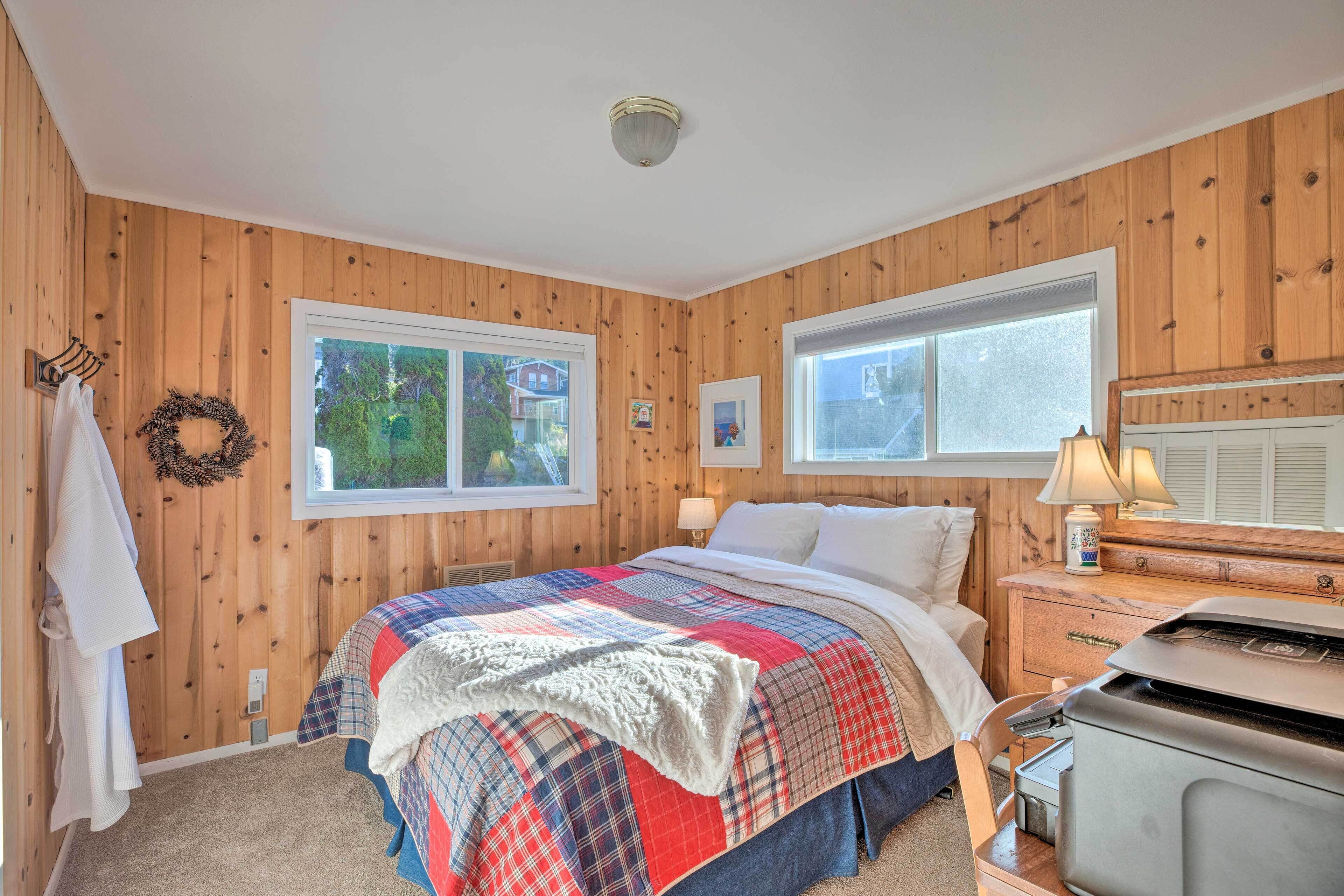 A cozy bedroom with wooden walls, a bed with a plaid comforter, a dresser, and a window with a view of greenery.