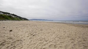 A wide sandy beach with a few scattered rocks, a grassy hill with buildings on the left, and a cloudy sky. - Exquisite Oceanside House w/ Pacific Views & Deck! (Tillamook)
