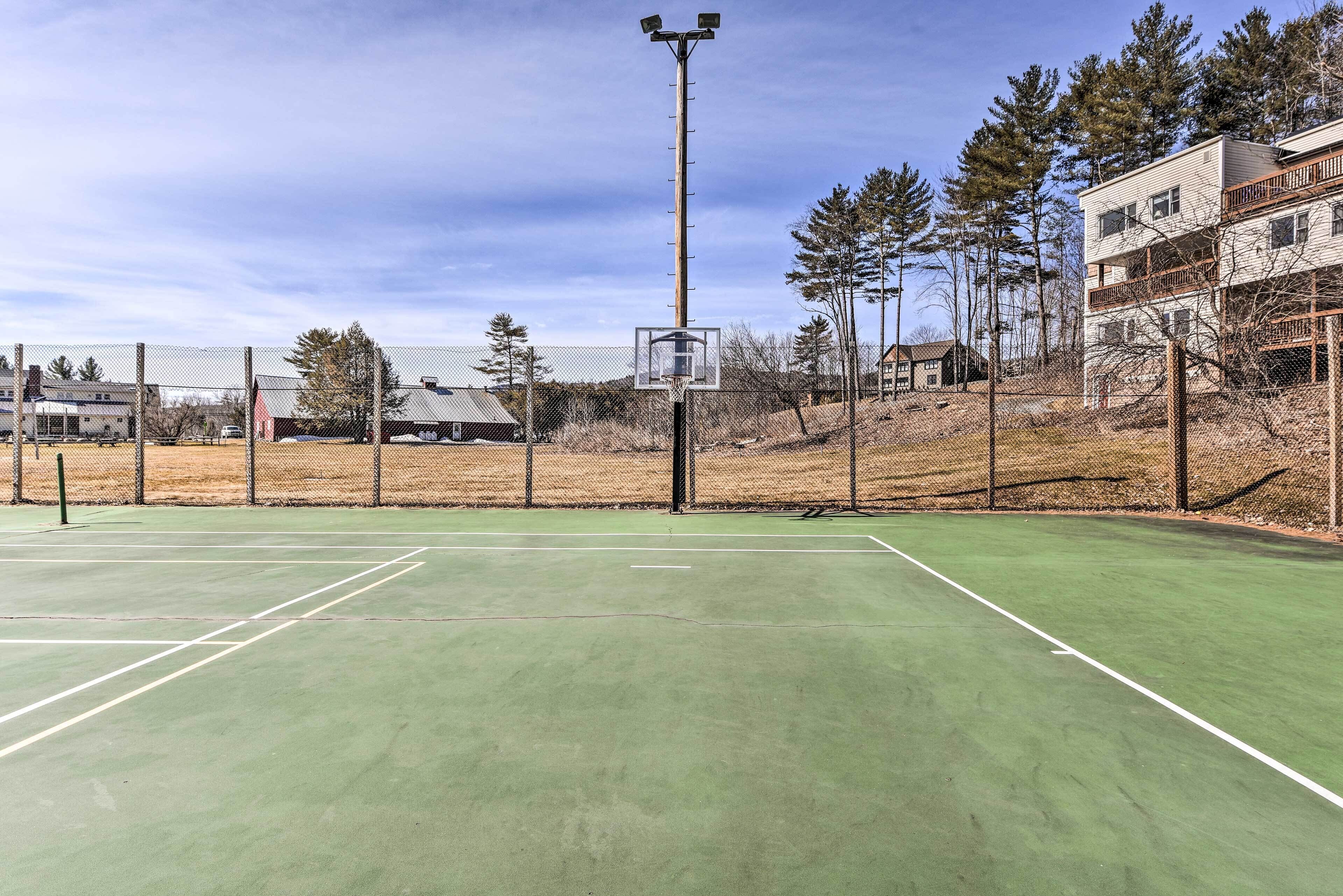 Una cancha de tenis al aire libre con un aro de baloncesto, rodeada por una cerca de malla, edificios y árboles.