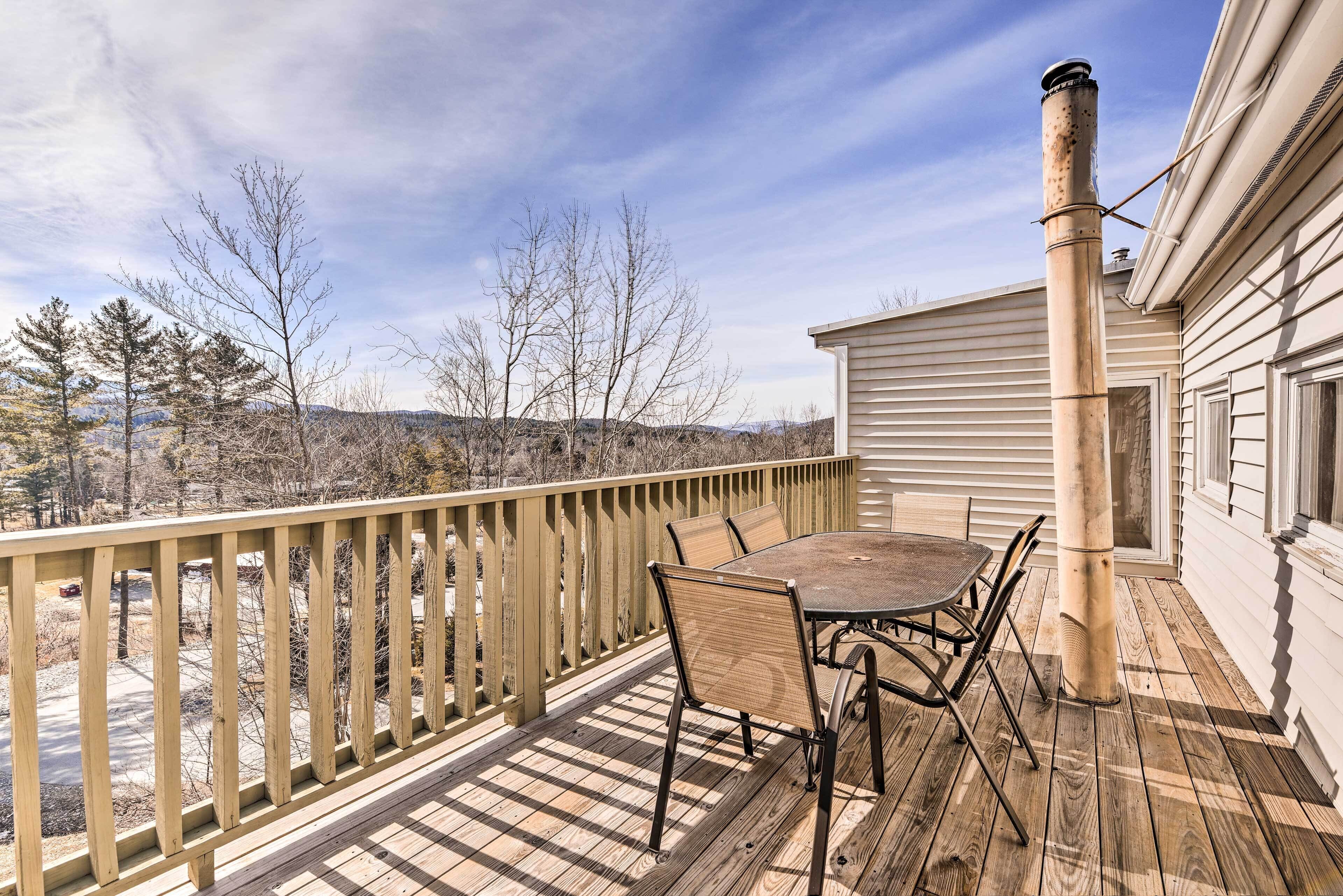 A wooden deck with a table and chairs, a chimney, and a view of trees and hills.