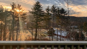 Blick vom Balkon auf eine bewaldete Landschaft, im Hintergrund ein Sonnenuntergang.
