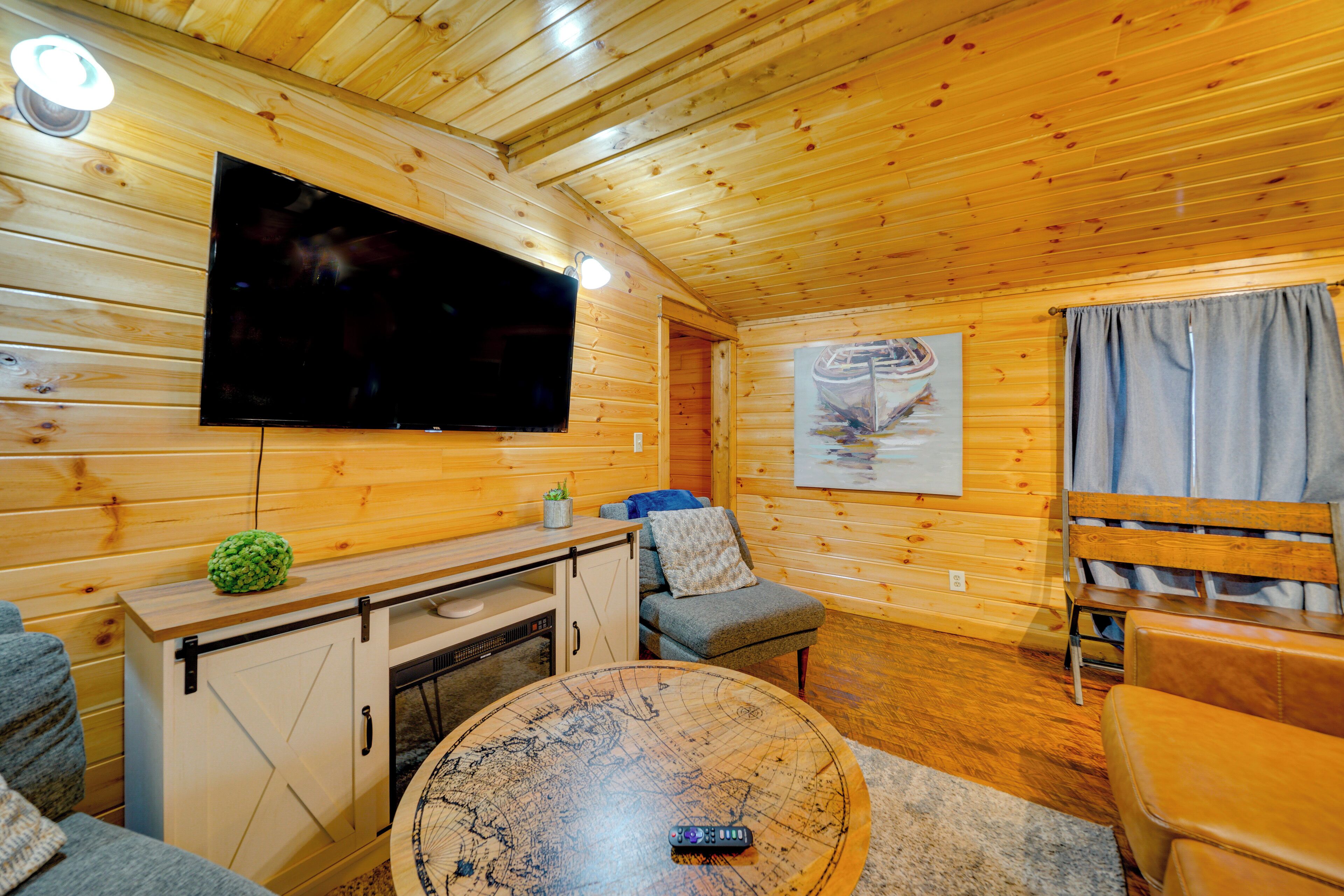 Interior acogedor de una cabaña con techo de madera, un televisor de pantalla plana en la pared, una mesa redonda con diseño de mapa, un sofá y un banco.