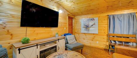 Interior acogedor de una cabaña con techo de madera, un televisor de pantalla plana en la pared, una mesa redonda con diseño de mapa, un sofá y un banco.