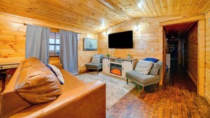 A cozy log cabin interior with a leather sofa, a fireplace, a television, and a small dining area. - Broken Bow Abode w/ Hot Tub < 7 Mi to Lake! (Broken Bow)