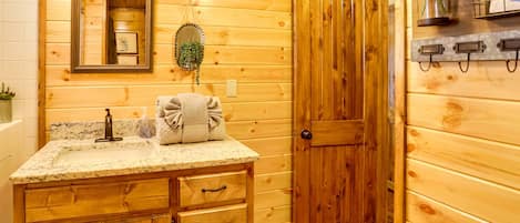 Un baño de cabaña de madera con lavamanos de encimera de granito, armarios de madera, un espejo, un perchero para toallas y una puerta.