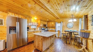 A wooden cabin interior with a kitchen featuring a stainless steel refrigerator, wooden cabinets, a central island with stools, and a dining table. - Broken Bow Abode w/ Hot Tub < 7 Mi to Lake! (Broken Bow)