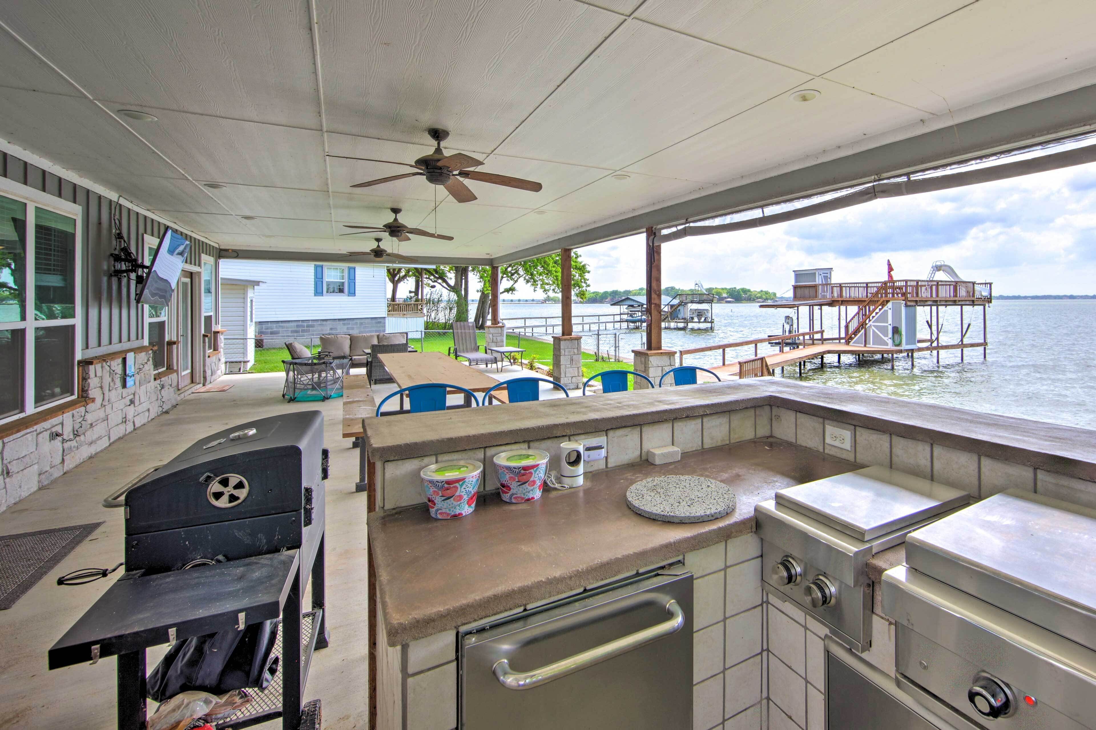 An outdoor kitchen area with a grill, a sink, and a countertop with containers. There are chairs and a table in the background, and a view of a dock and water.