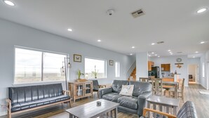 Un vaste espace de vie comprenant un canapé, une table basse et une table à manger. Au fond, on aperçoit une cuisine avec des meubles en bois et un escalier menant à l’étage supérieur.