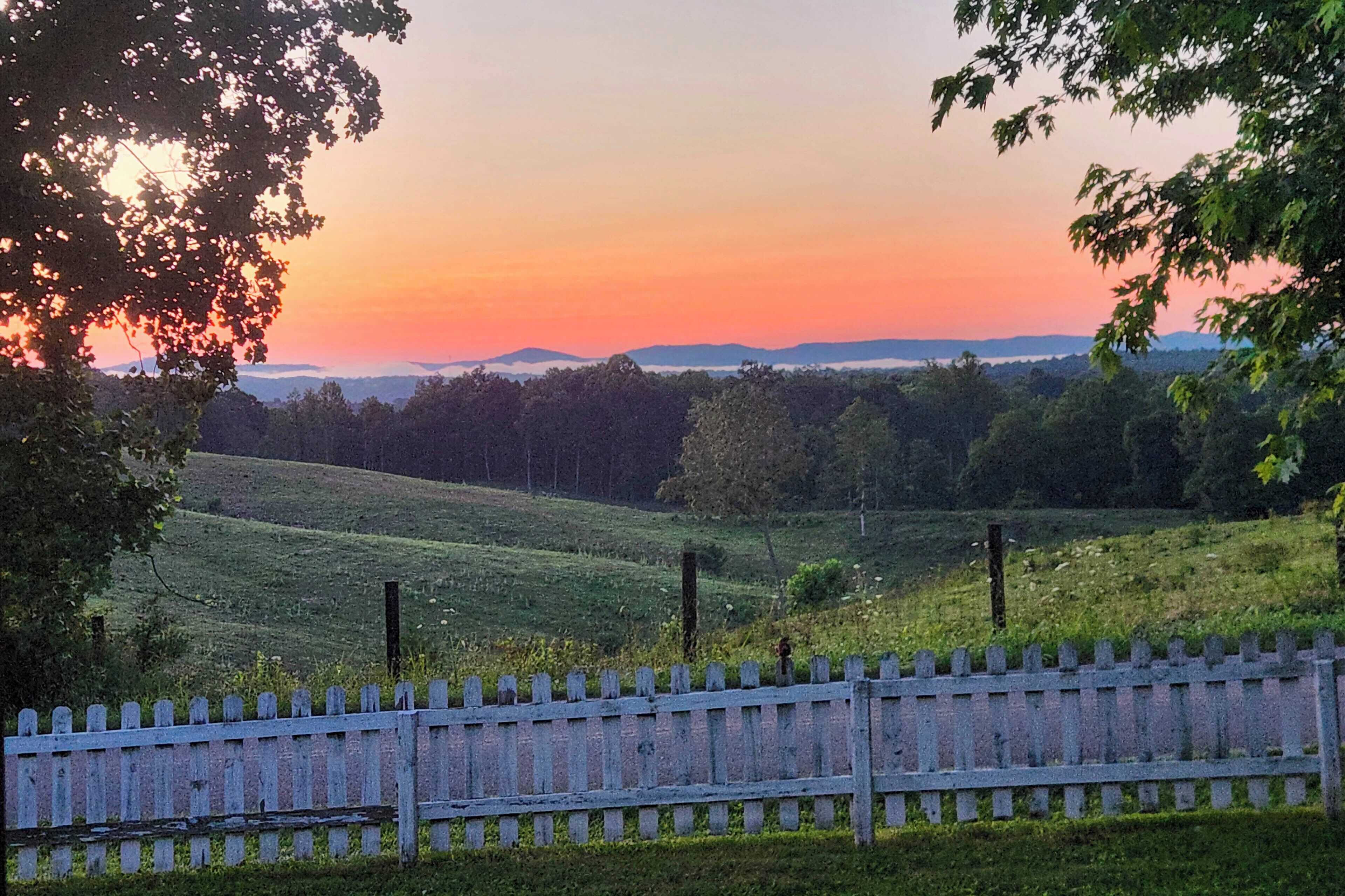 A rural landscape with a wooden fence, grassy fields, and a sunset in the background.