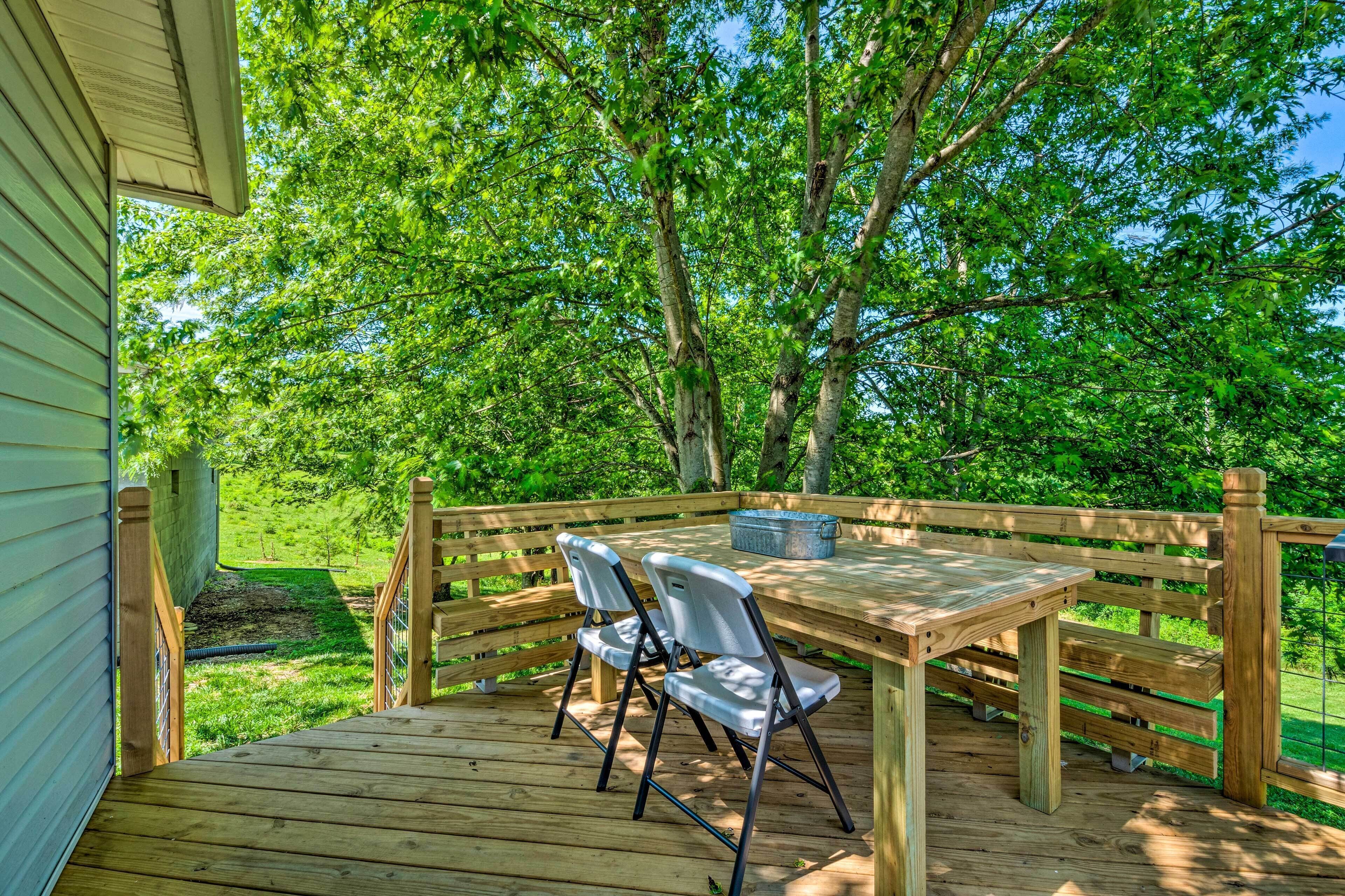 A wooden deck with a table and two chairs, surrounded by a wooden fence and lush greenery.