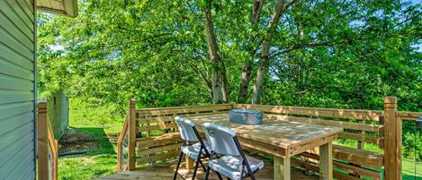 A wooden deck with a table and two chairs, surrounded by a wooden fence and lush greenery.