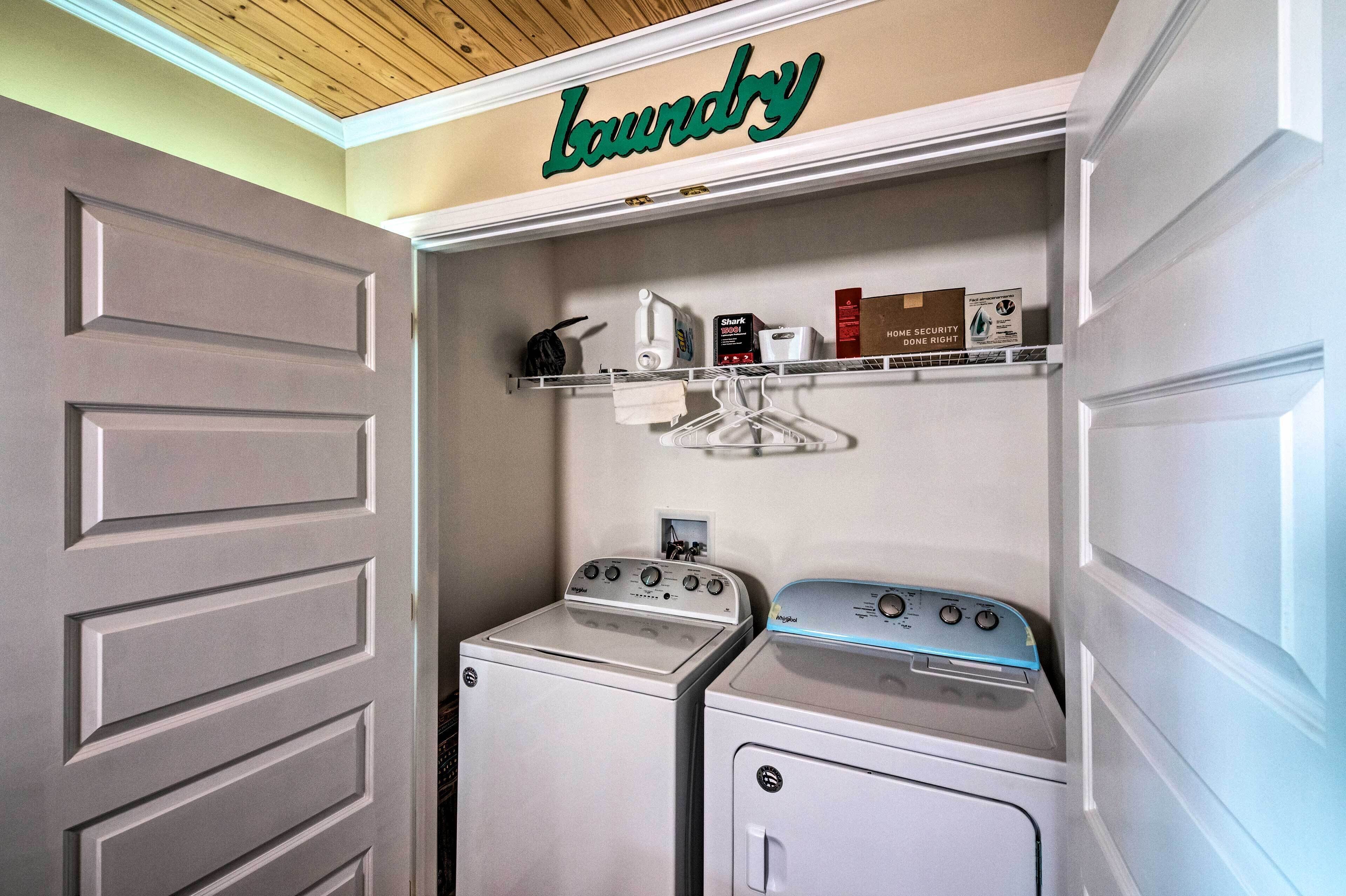 A laundry room with a 'Laundry' sign, two washing machines, a dryer, and a shelf with various items.
