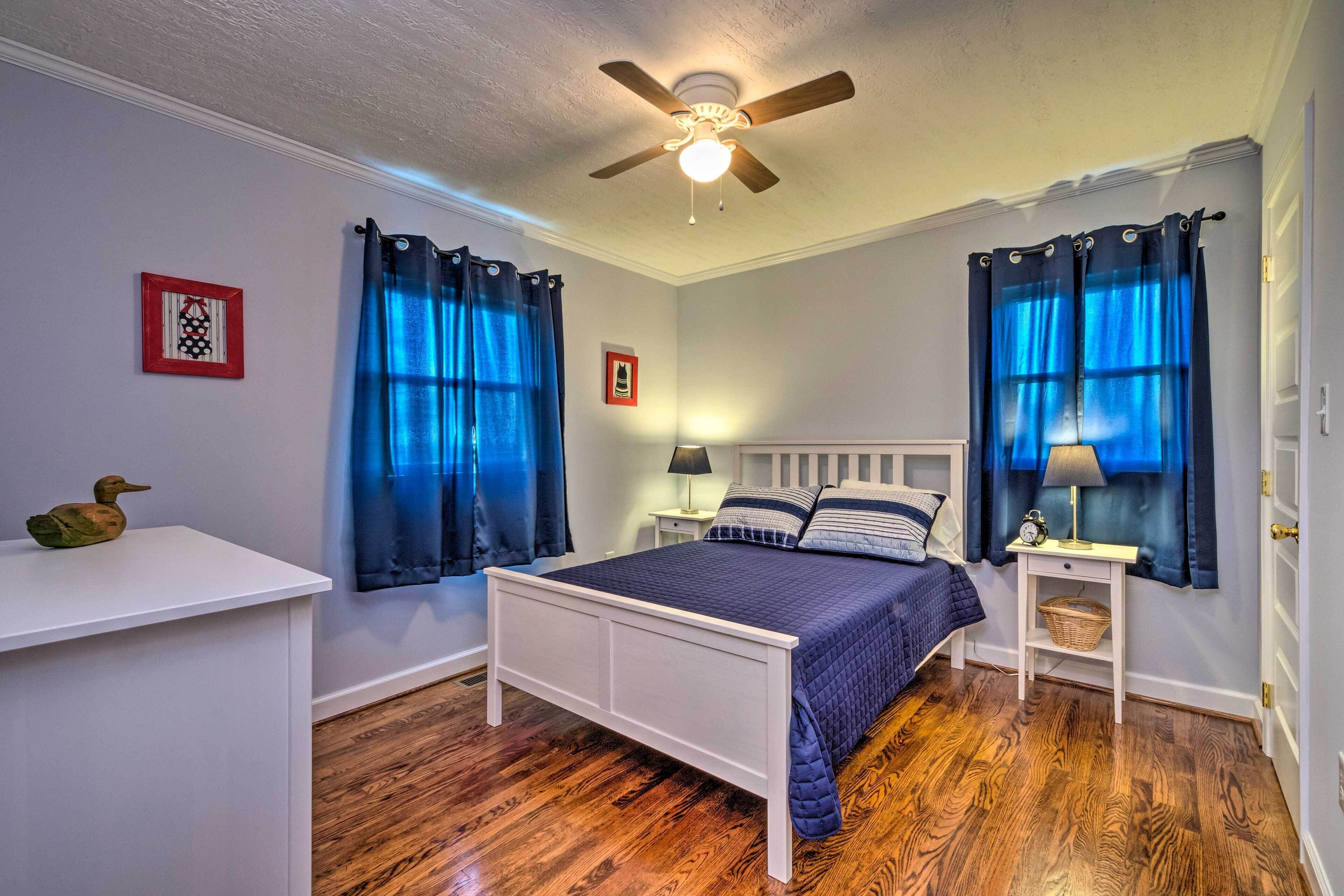 A bedroom with a white bed, blue curtains, a ceiling fan, and wooden flooring.