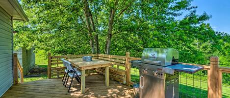Una terraza de madera con una parrilla, una mesa y sillas, rodeada de árboles.