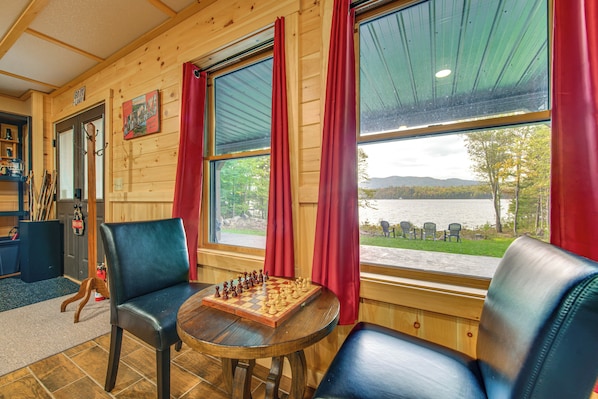 Un interior de madera con un tablero de ajedrez sobre una mesa, dos sillas, vista a un lago y una puerta con un letrero.