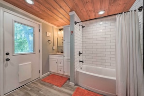 A bathroom with a white bathtub, a shower with a white curtain, a window, and a wooden ceiling.