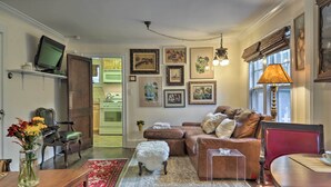 A cozy living room with a sofa, ottoman, and a small table. There is a television on a shelf, a lamp, and various framed pictures on the wall. - Historic Ocean Grove Home - Walk to Beach! (Ocean Grove)