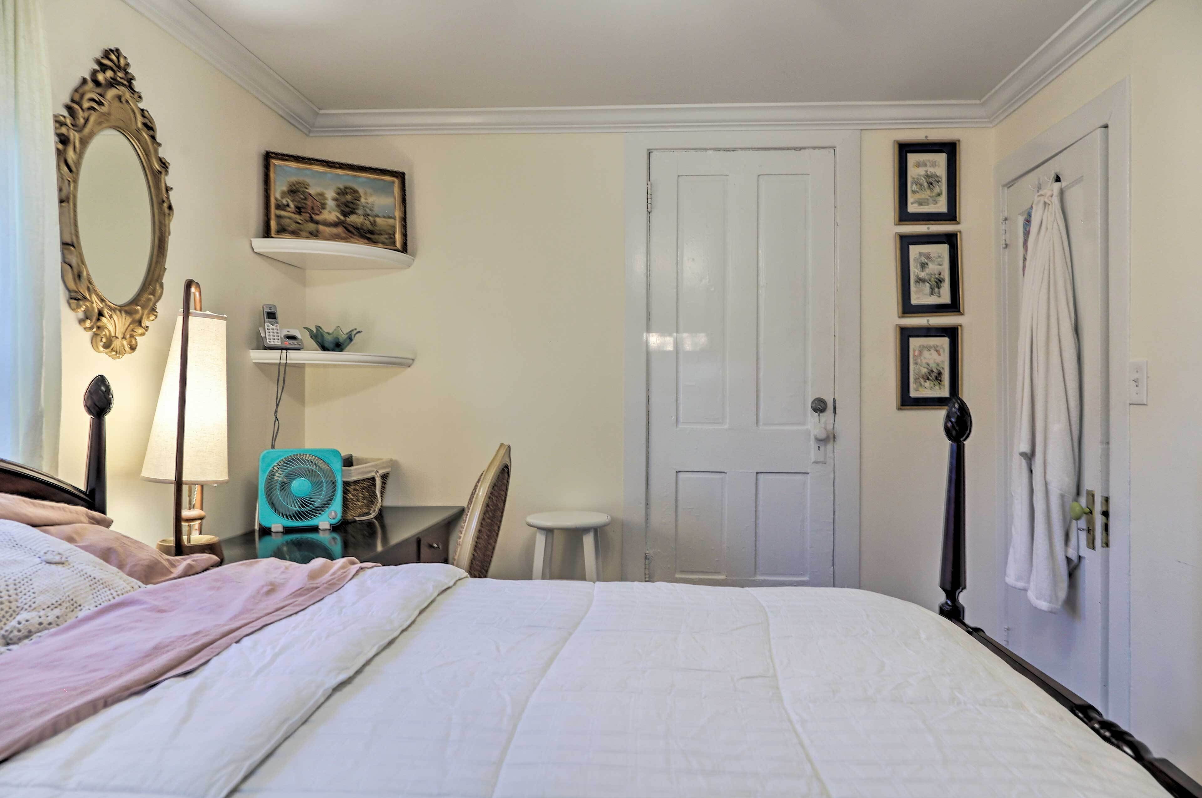 A bedroom with a bed, a bedside table with a lamp, a fan, a chair, a door, and a wall with framed pictures.