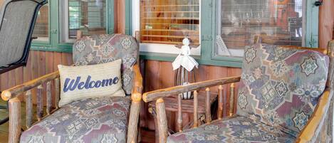 Two patterned armchairs with a 'Welcome' pillow, a wooden chair, and a window with blinds.