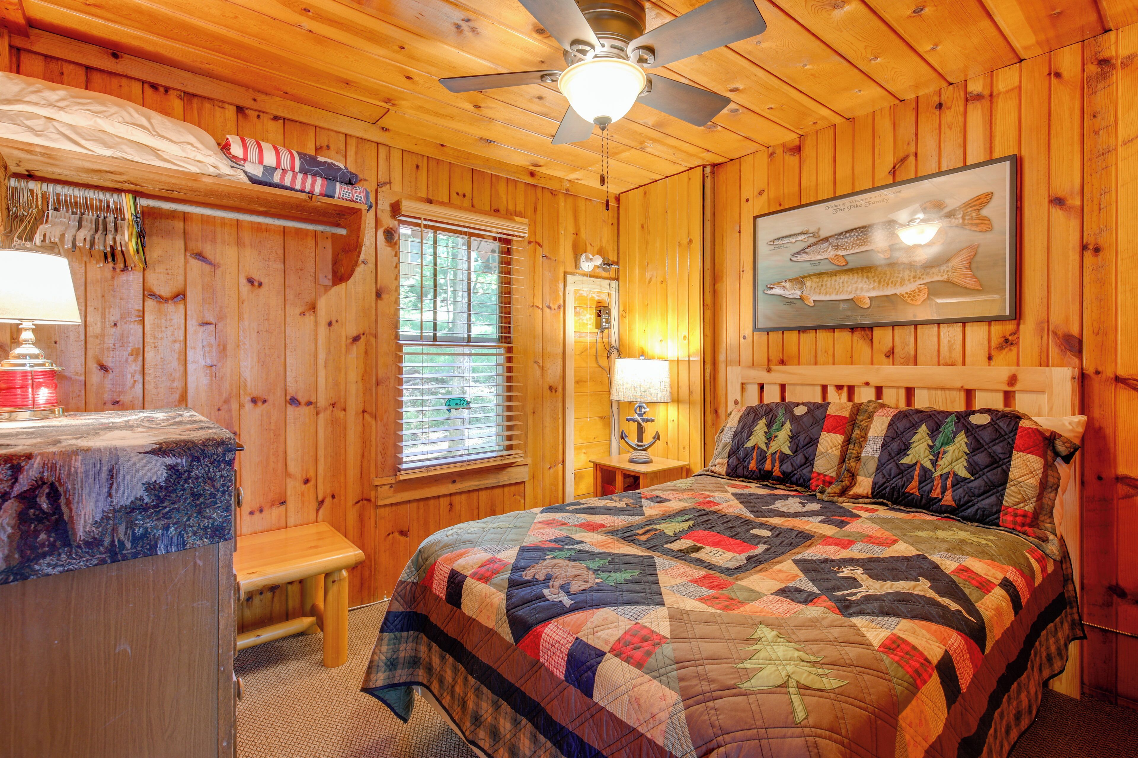 A wooden cabin bedroom with a bed, a ceiling fan, a window with blinds, a nightstand with a lamp, and a framed fish picture on the wall.