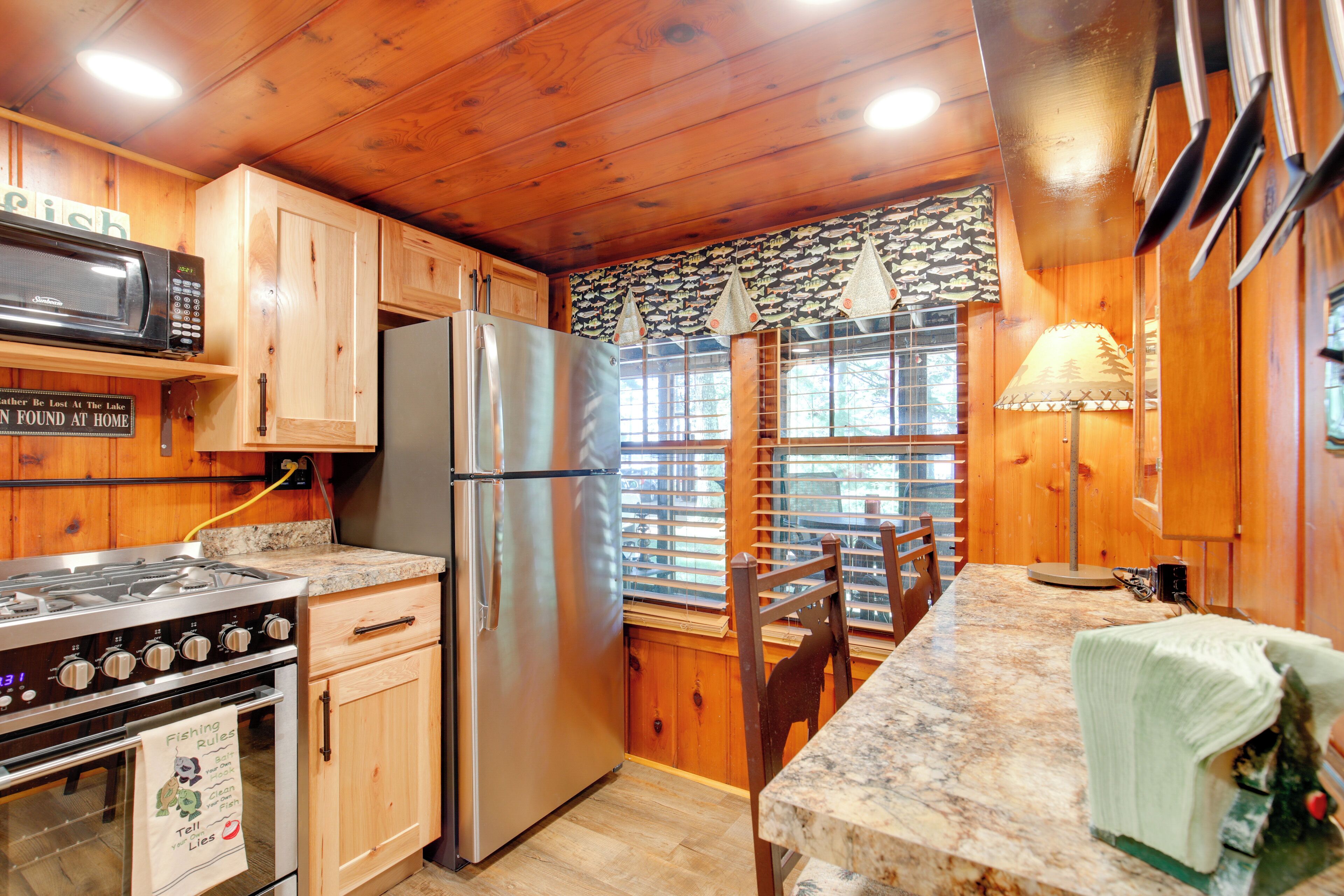 A kitchen with wooden cabinets, a stainless steel refrigerator, a microwave, a stove, and a window with blinds.