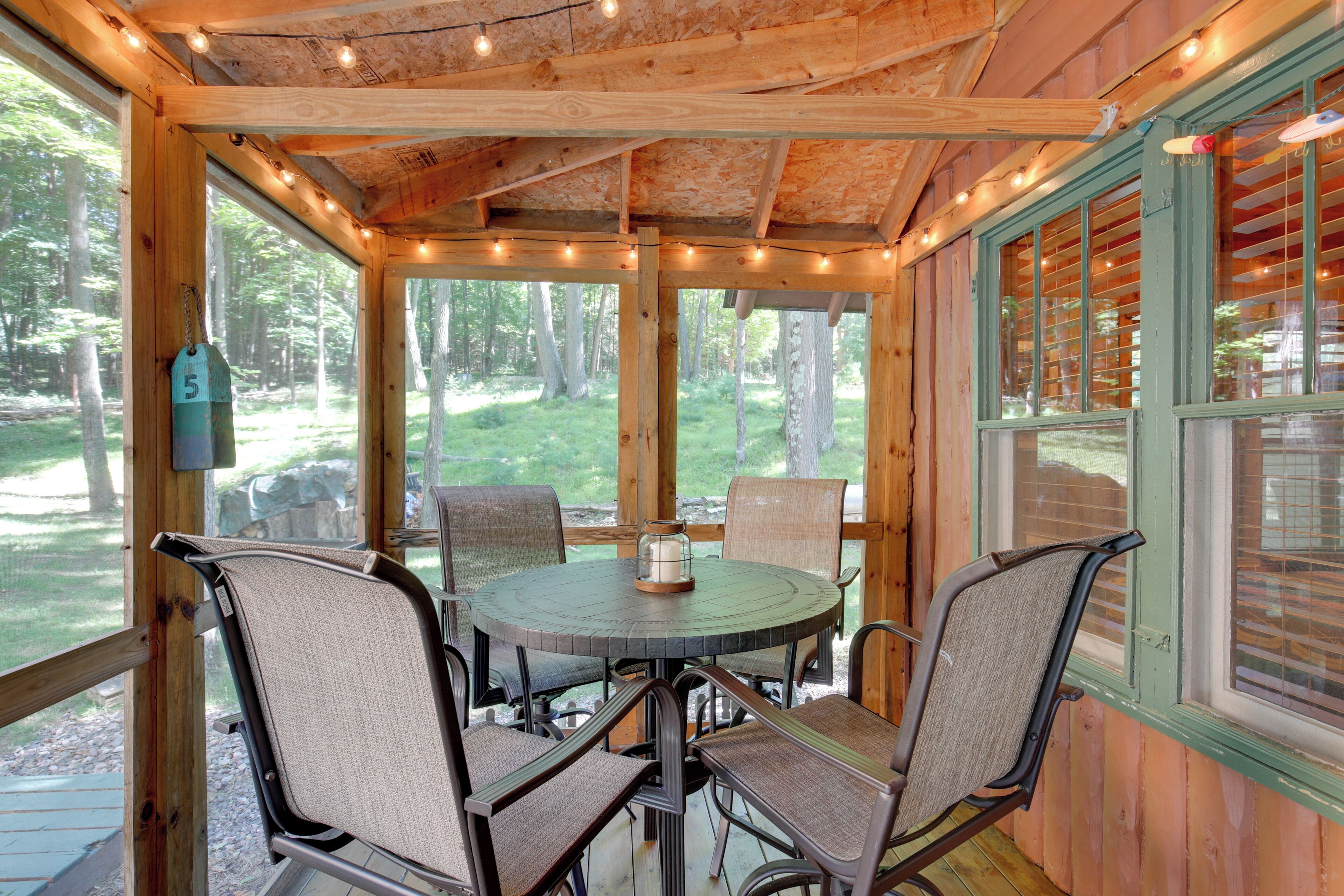 A wooden cabin with a round table and four chairs, a hanging tag, and a view of the forest through large windows.