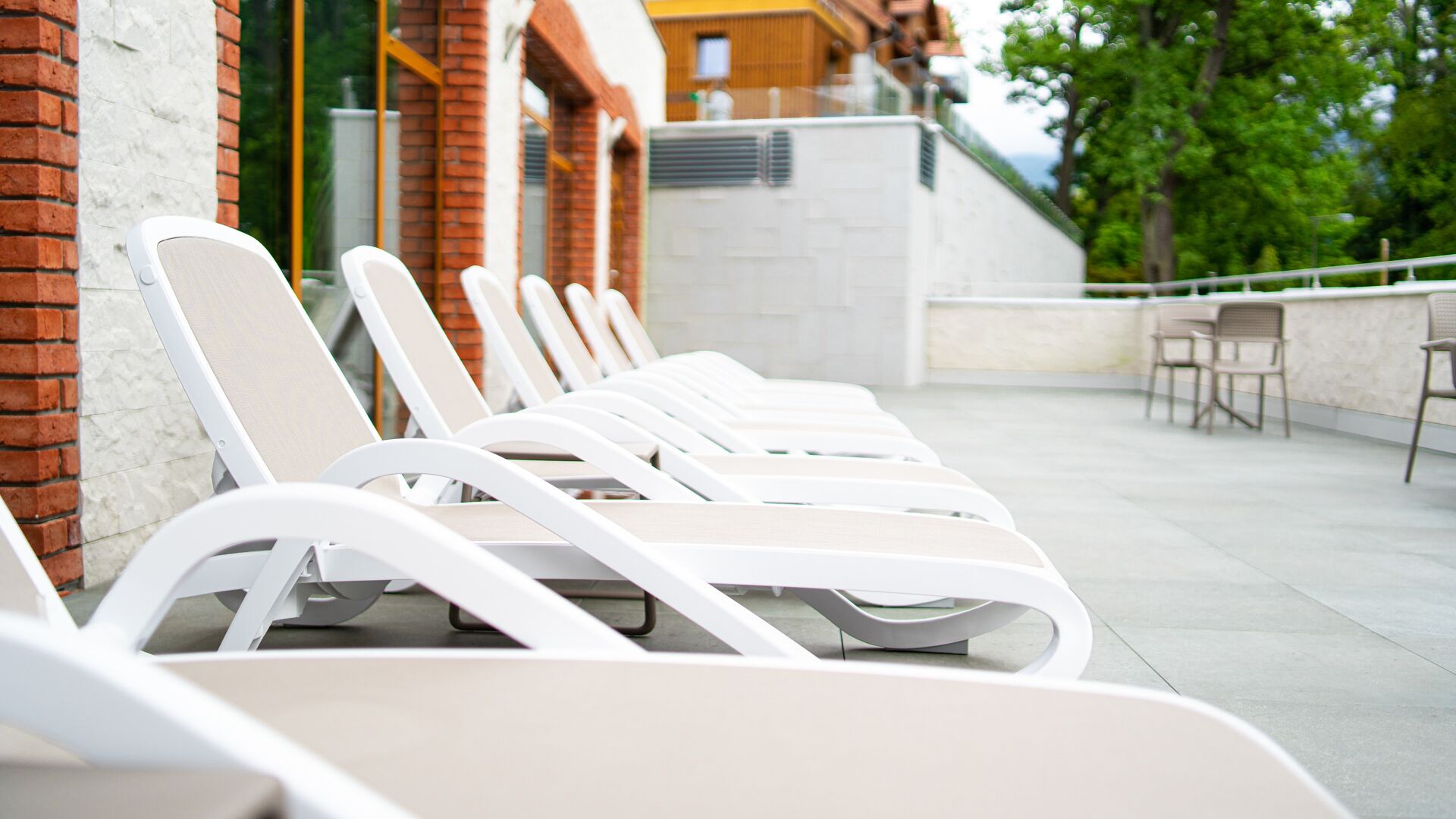 A row of white lounge chairs aligned against a brick wall with a white ceiling and a view of trees and a building in the background.