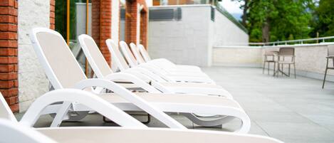 A row of white lounge chairs aligned against a brick wall with a white ceiling and a view of trees and a building in the background.
