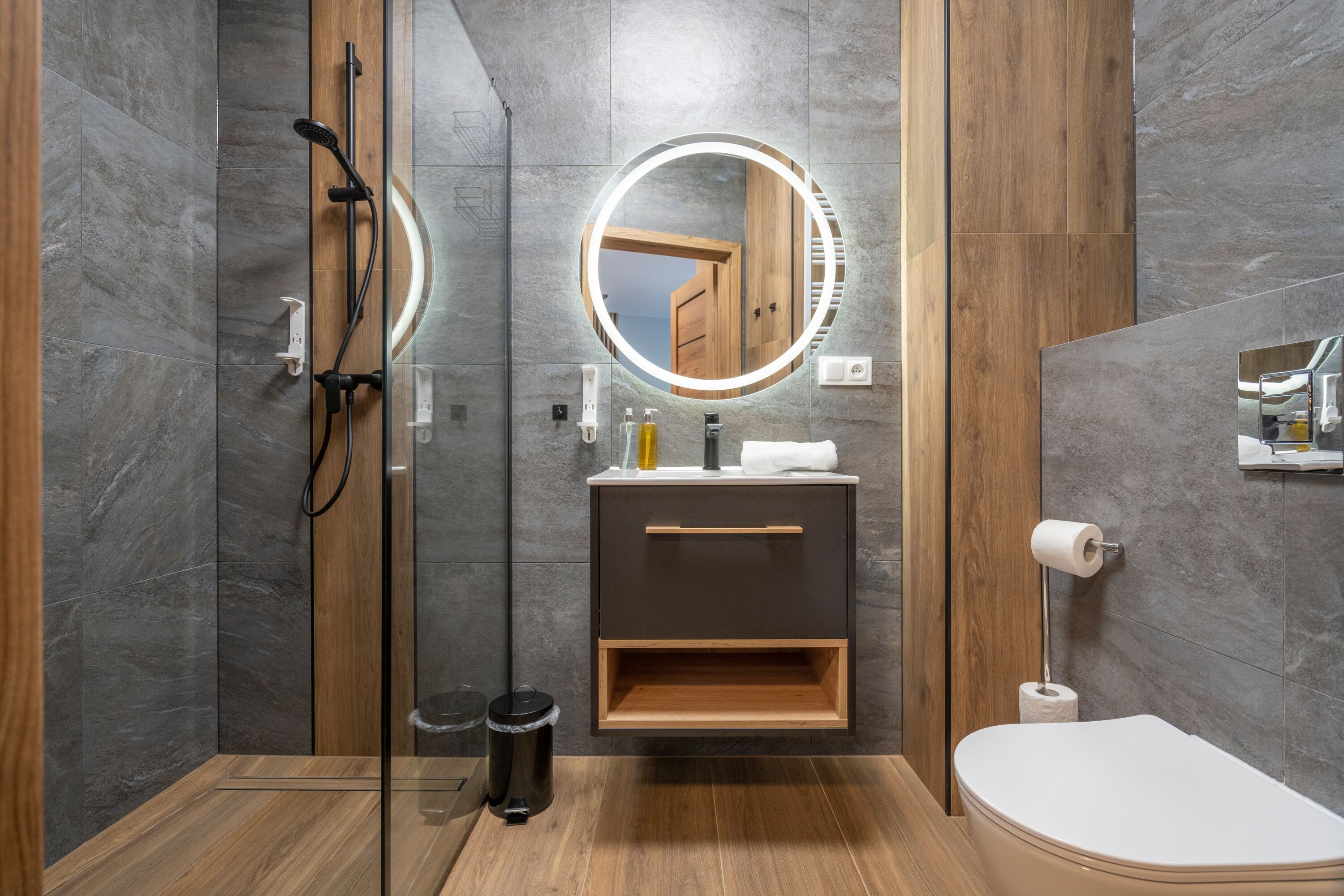 Een moderne badkamer met een glazen douchecabine, een houten wastafelmeubel, een toilet en een ronde spiegel met verlichting.