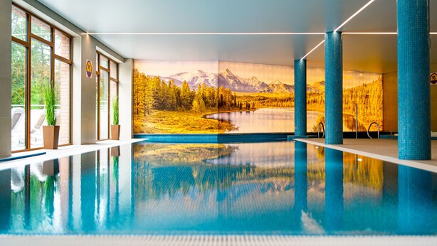 An indoor swimming pool with a mountain landscape mural on the wall, large windows, and blue pillars. - RentPlanet - Stone Hill (Szklarska Poreba)
