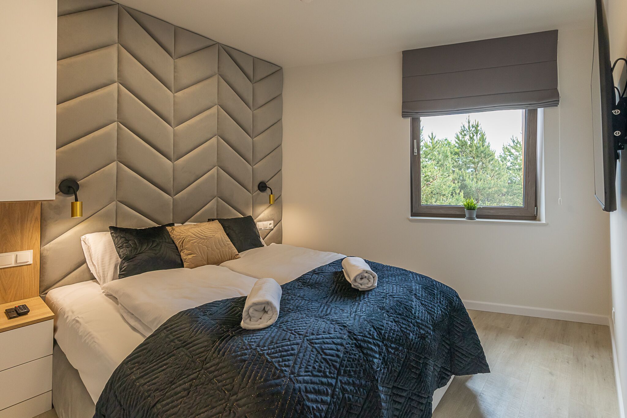 Une chambre moderne avec un grand lit, une tête de lit ornée d’un motif géométrique, une table de chevet avec une lampe et une fenêtre donnant sur des arbres.